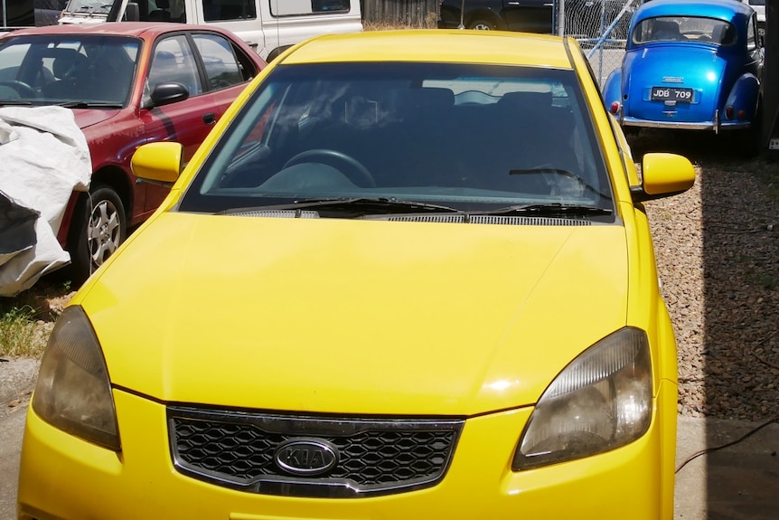 A yellow Kia Rio, a white Defender and a blue Morris Minor in a car yard.