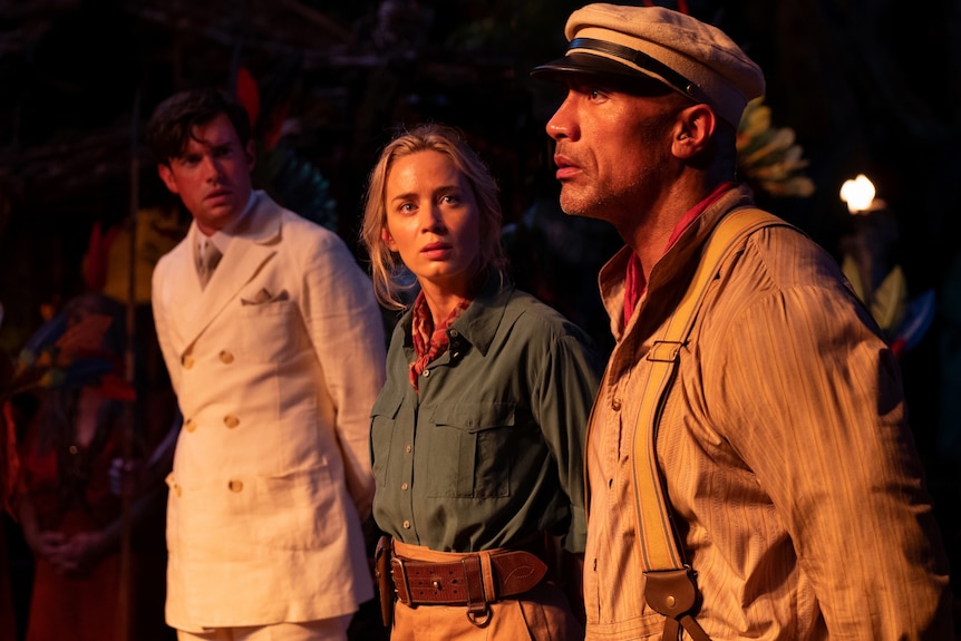 Lit by torchlight, a dishevelled Jack Whitehall and Emily Blunt look over in confusion at Dwayne Johnson, who seems concerned