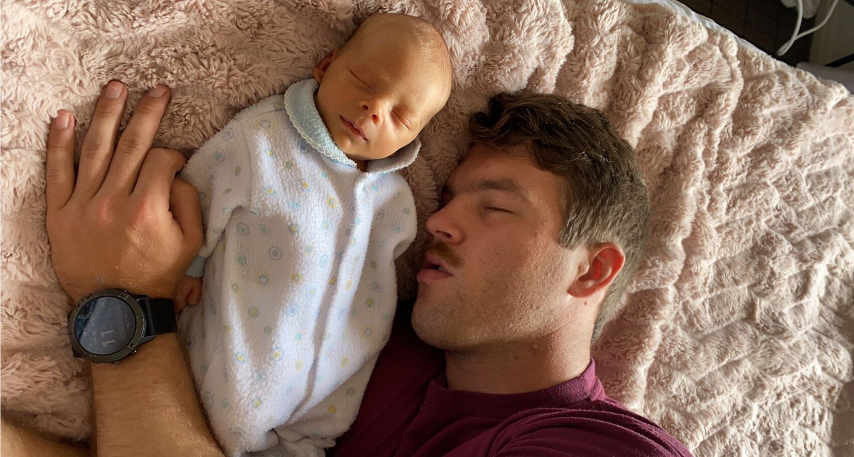 A man asleep on a bed beside a sleeping newborn, with his arm around the baby.