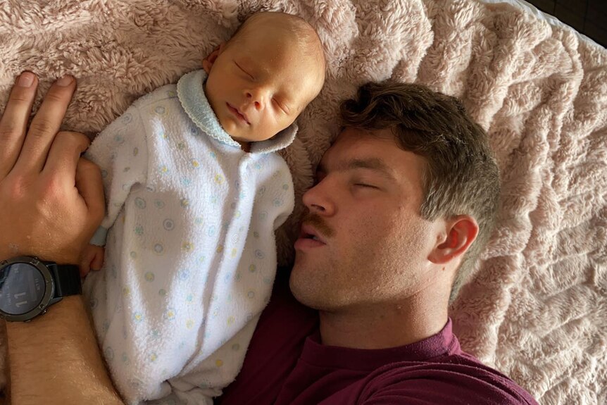 A man asleep on a bed beside a sleeping newborn, with his arm around the baby.