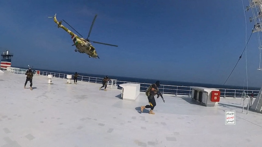 A helicopter flies over a large ship with four gunmen with covered faces running on deck