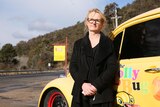 Woman in a black coat and glasses standing infront of a bright yellow car with a 'Lolly Bug' sign in the background