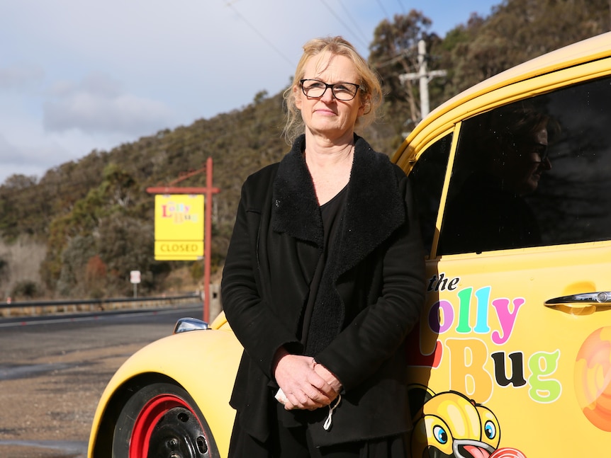 Woman in a black coat and glasses standing infront of a bright yellow car with a 'Lolly Bug' sign in the background