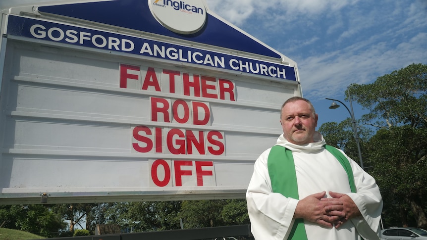 Priest stands out the front of a church sign which reads father rod signs off 