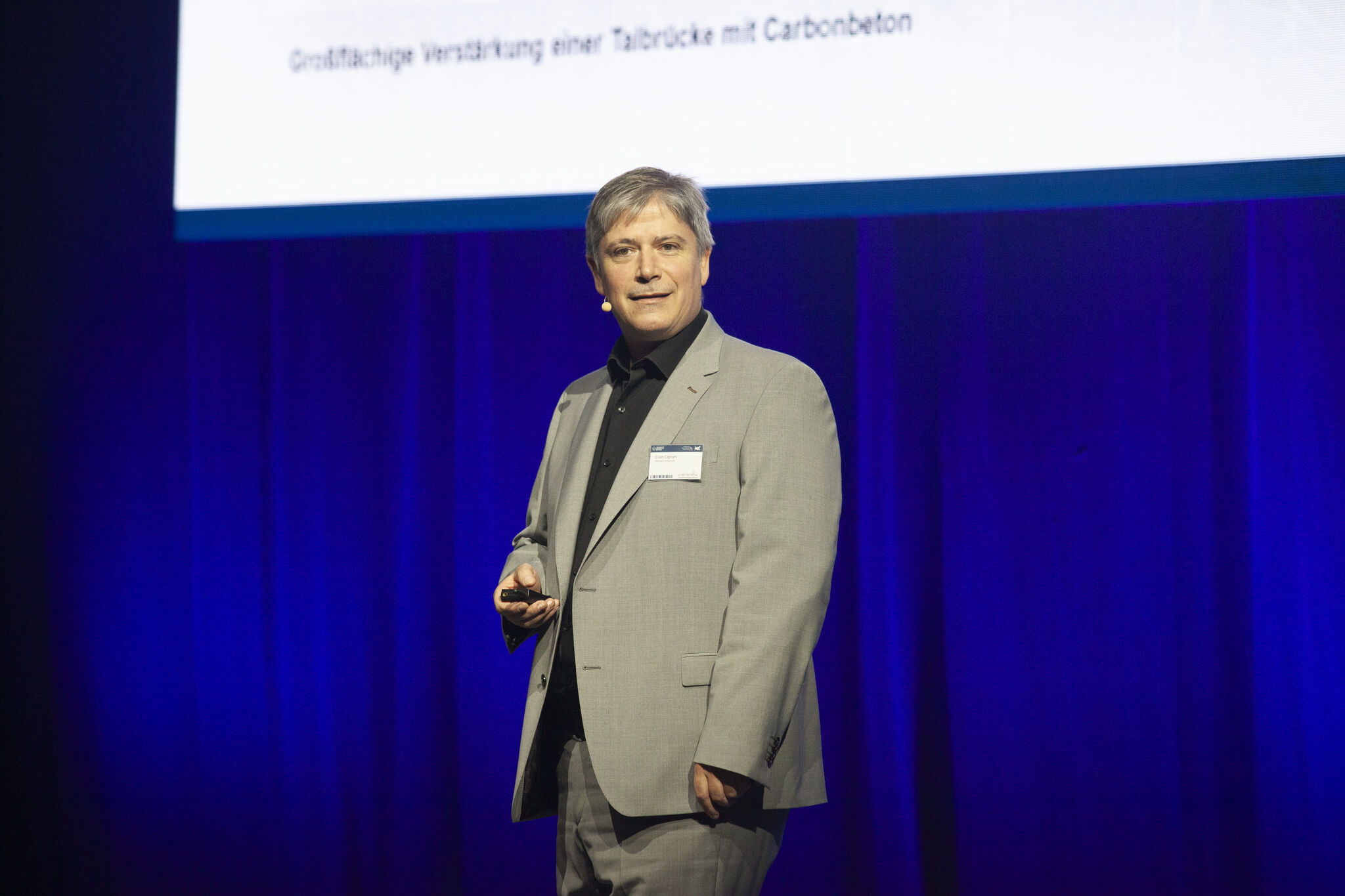 Colin Caprani está en un escenario frente a un telón azul y viste un traje beige.