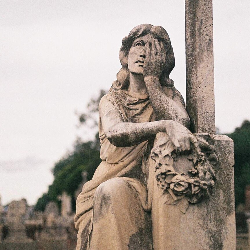 You view a statue of a woman learning against a tombstone on an overcast day in a dense cemetery.