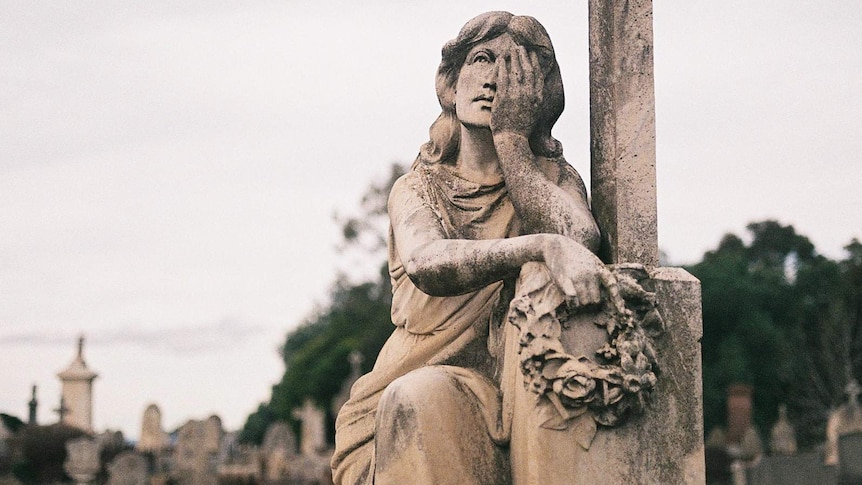 You view a statue of a woman learning against a tombstone on an overcast day in a dense cemetery.