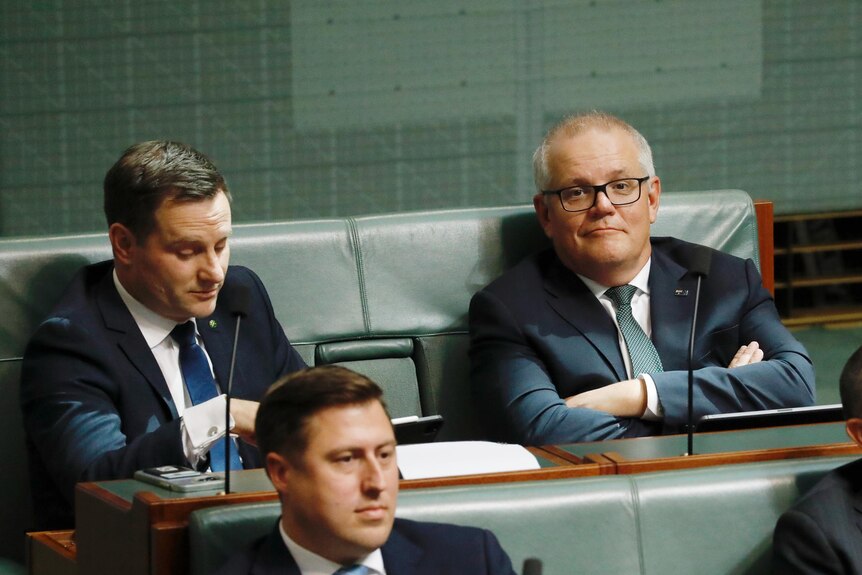 Morrison sits with his arms folded in his seat in the house of representatives, looking at the camera, hawke beside him.