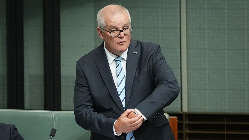 Morrison holds up his wrist showing a plastic bracelet, standing in the lower house chamber.