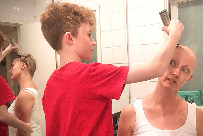 A young boy in a red tshirt shaves a woman's head at home.