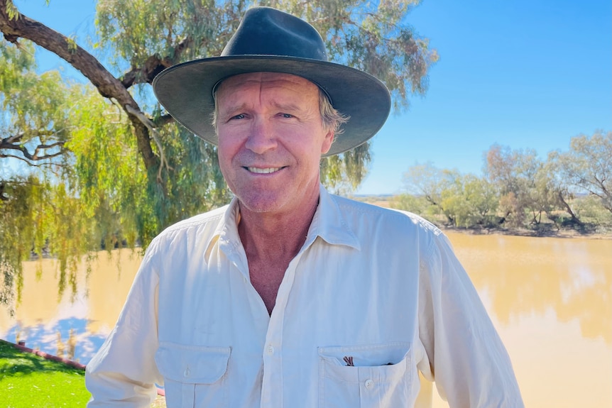 A man looks at the camera smiling, wearing a grey hat. He stands in front of trees and a small lake