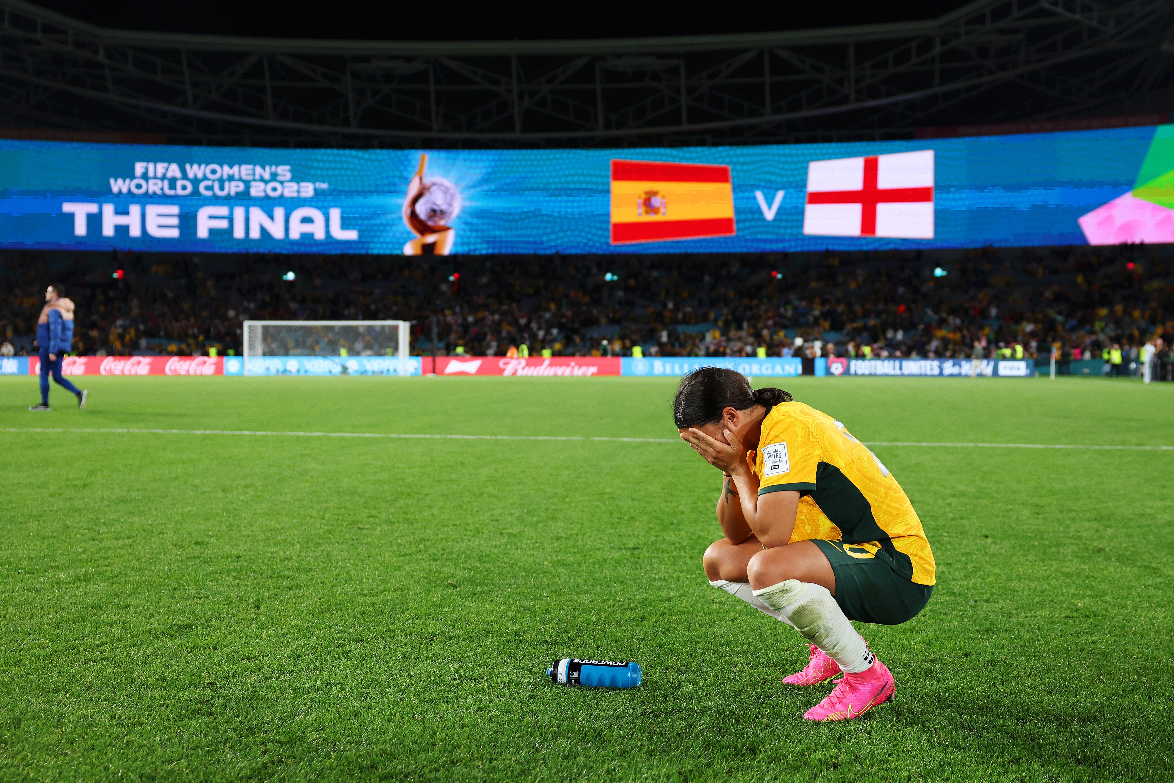 A soccer player wearing yellow and green holds her face in her hands with the result on the scoreboard behind