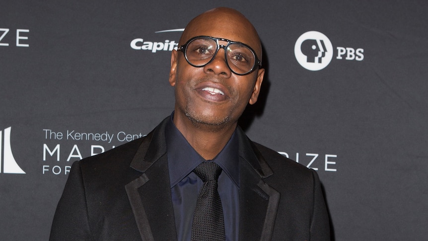 Dave Chappelle looks in the direction of the camera, wearing a suit and dark-rimmed glasses. 