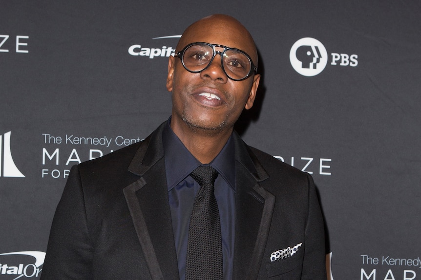 Dave Chappelle looks in the direction of the camera, wearing a suit and dark-rimmed glasses. 