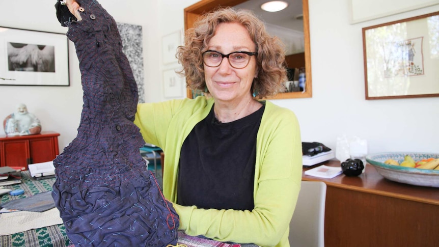 A woman holds up a piece of embroidered and pleated textile art as she sits at a table inside an art studio.