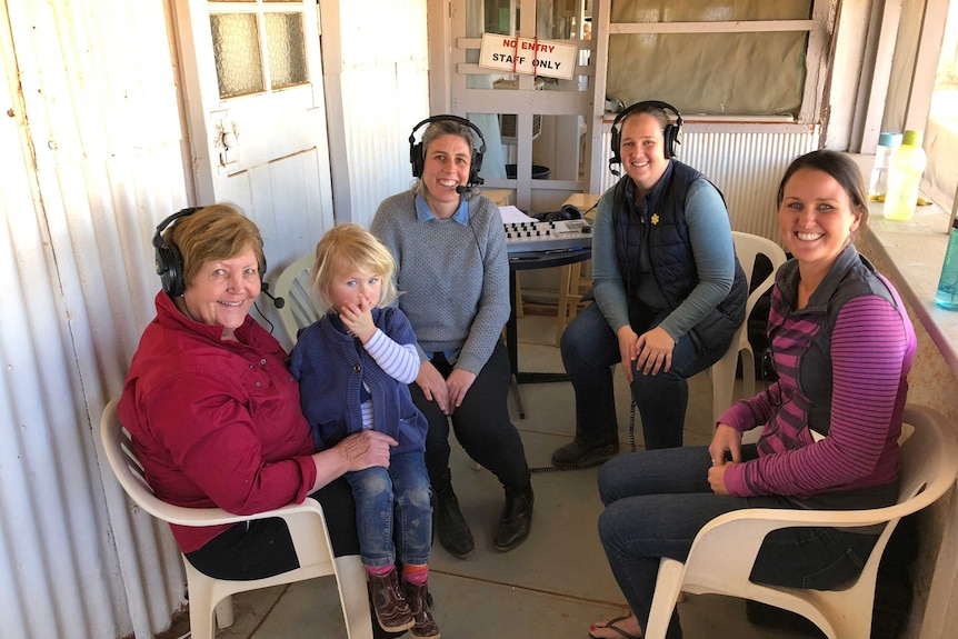 Neindorf with a group of women and children wearing headphones sitting on verandah.