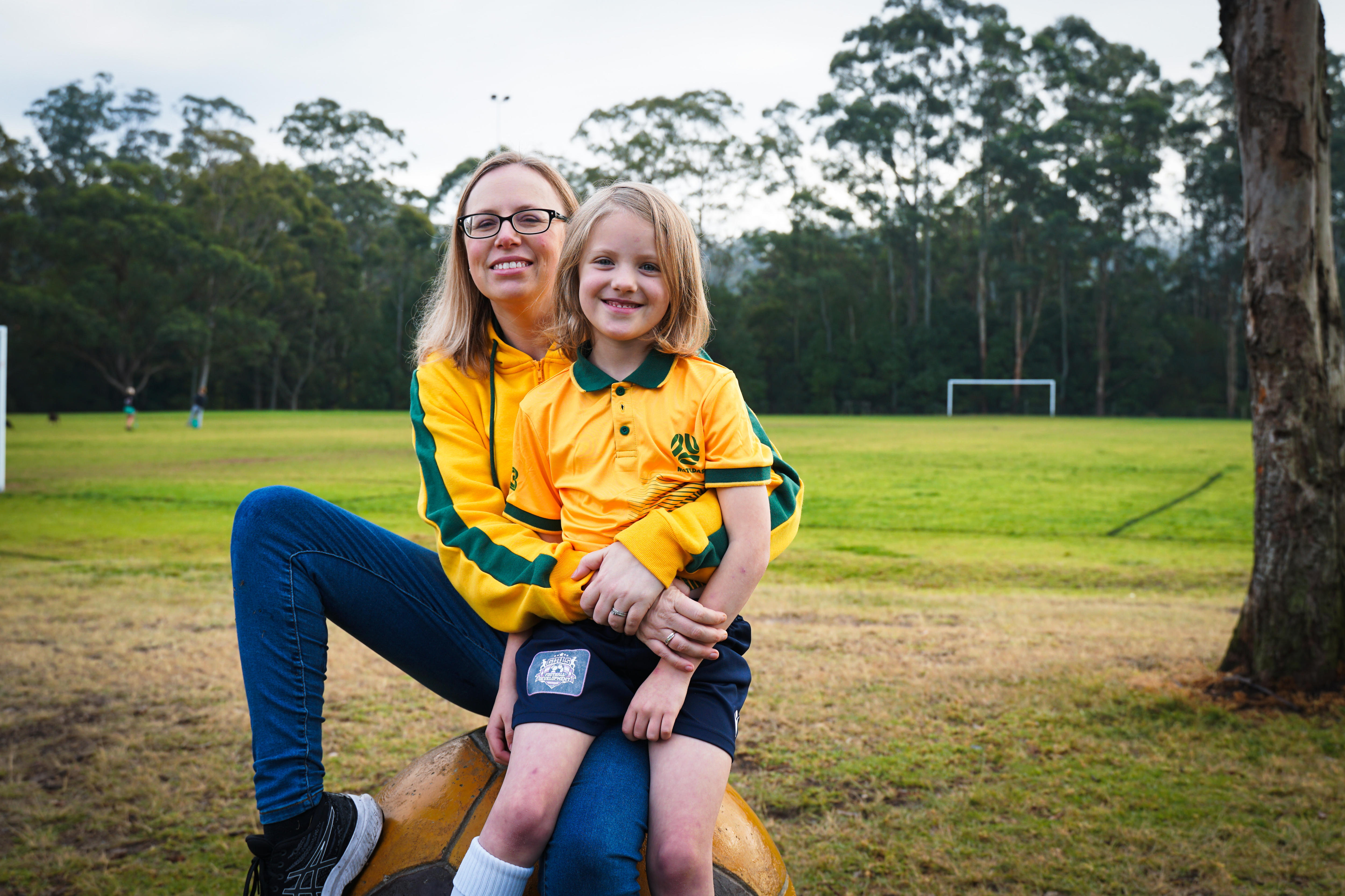 A woman wearing a bright yellow jacket sits on an oversized soccer ball, a child sits on her lap. They're on a football field.
