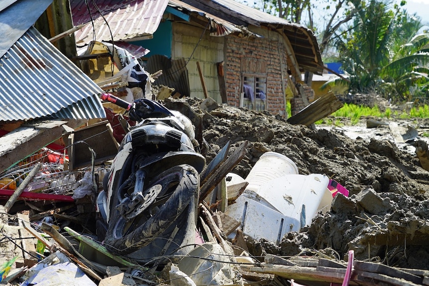 A motorbike is half-covered by debris and rubbish next to a huge pile of mud and a damaged house