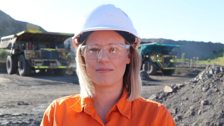 Close-up shot of woman wearing hi-vis, hard-hard and safety glasses, with two coal trucks in background.