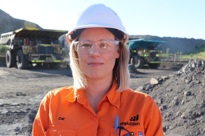 Close-up shot of woman wearing hi-vis, hard-hard and safety glasses, with two coal trucks in background.