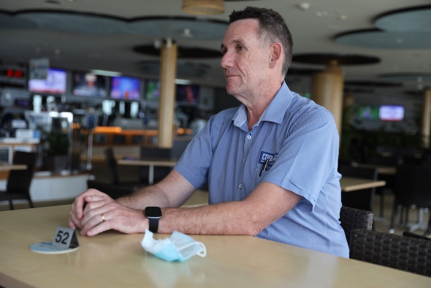 Man in blue checkered shirt, short hair, sitting at a table in a surf club, tvs and bar in background