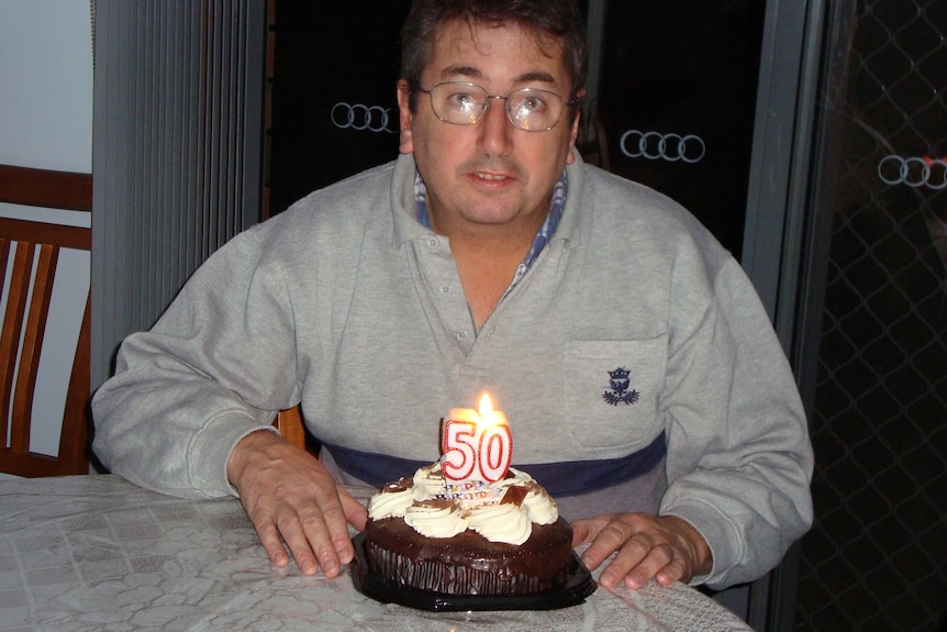 A man wearing a grey jumper with a birthday cake with the number 50 and a candle on top