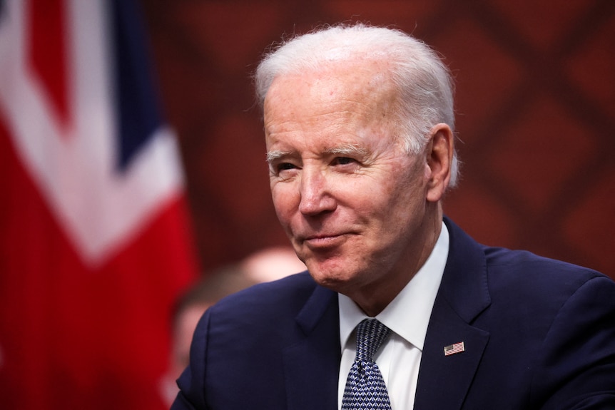 Joe Biden smiles while sitting infront of a US flag in a conference room.