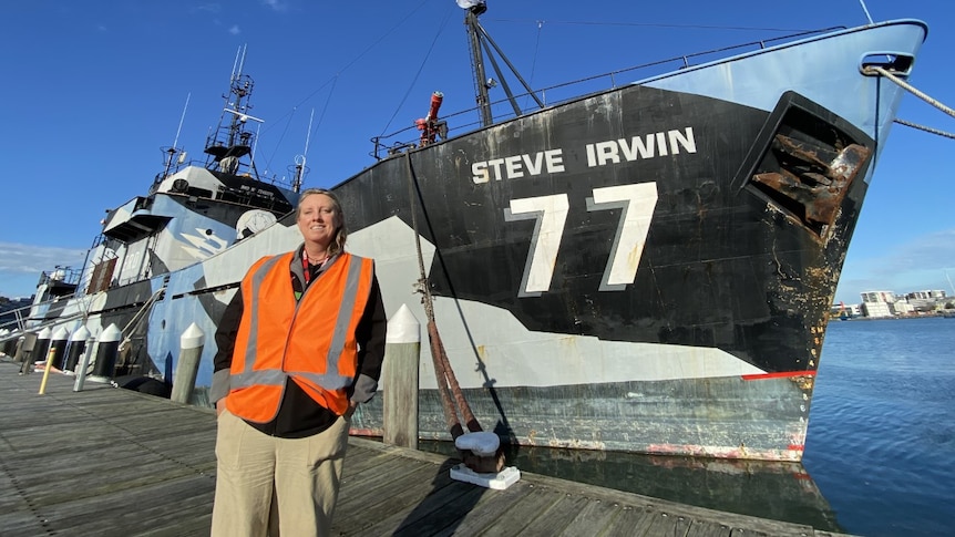 A woman in a orange vest standing in front of a ship that has "Steve Irwin 77" written on it.