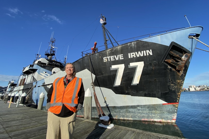 A woman in a orange vest standing in front of a ship that has Steve Irwin 77 written on it