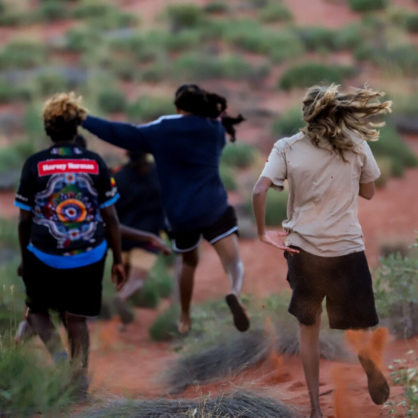 Four young Aboriginal women run down a red sand dune barefooted 