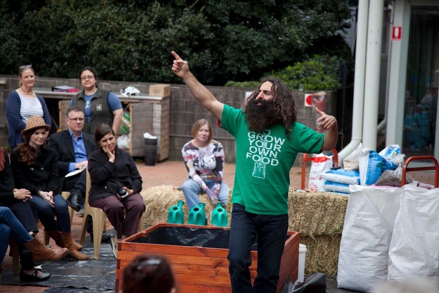 Costa in green t-shirt with grown your on food written on it pointing to sky as people sitting on hay bales watch on.