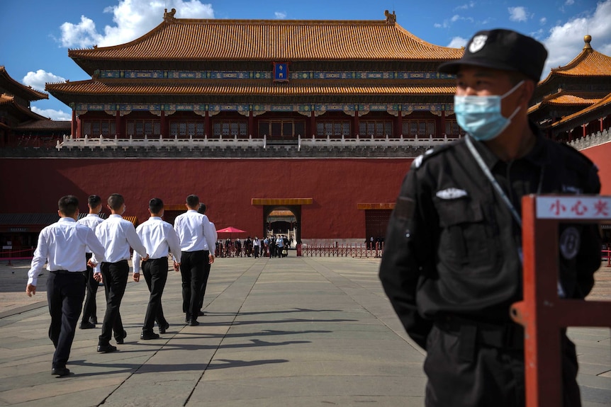 A male security guard wearing a face mask stands with hands clasped behind back while a group of six men march past.