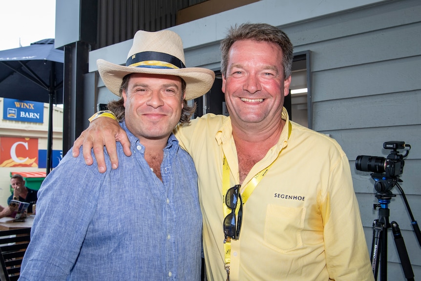 A man in a yellow shirt stands smiling with his arm around another man, in a blue shirt and hat.