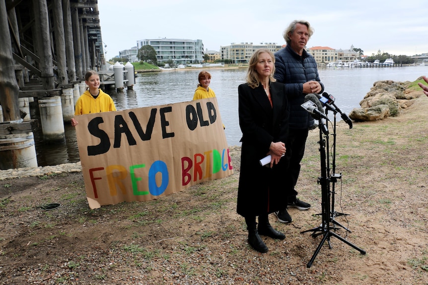 A man and a woman stand on the shore of the Swan River at a microphone with two children holding a banner behind them.