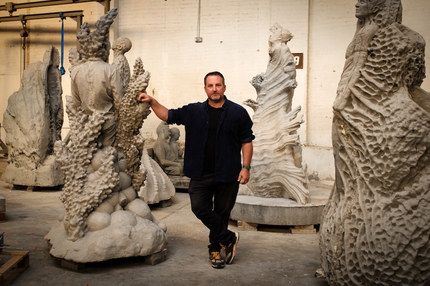 A man poses with large concrete sculptures that are half-human, half-marine life