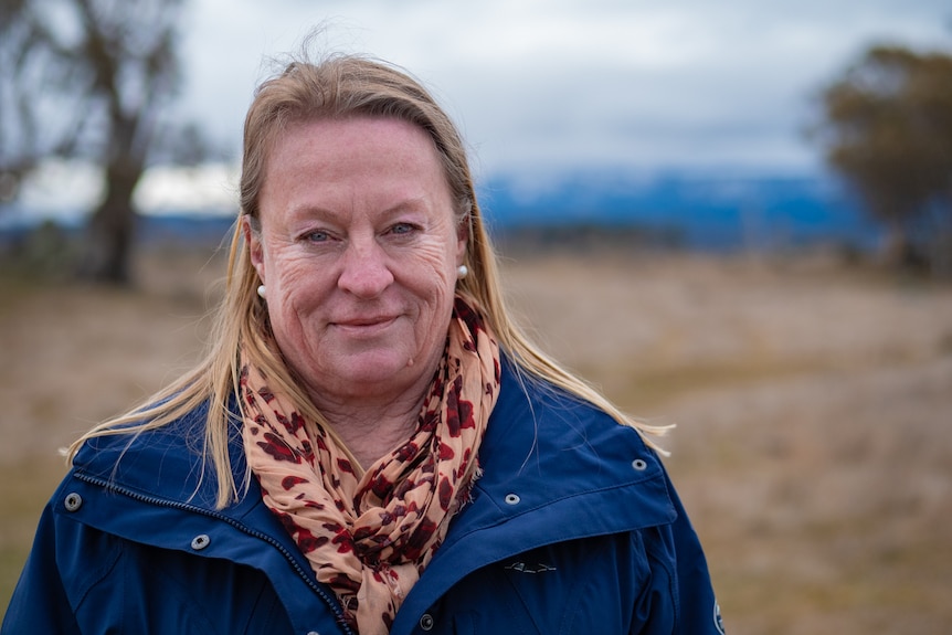 A woman with long blonde hair, blue jacket and coral spotted scarf stares at the camera, mountains behind her.