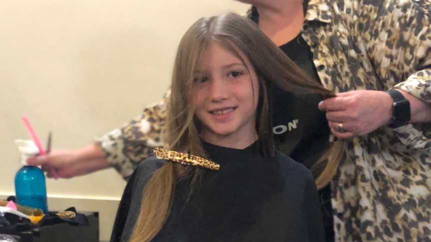 A young girl with long light brown hair is getting her hair cut. The hairdresser behind her reaches for the spray bottle.