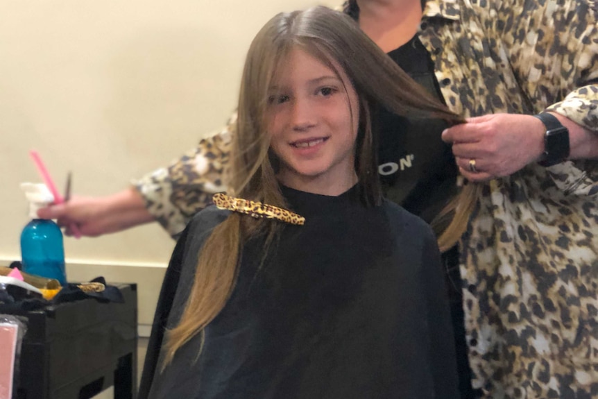 A young girl with long light brown hair is getting her hair cut. The hairdresser behind her reaches for the spray bottle.