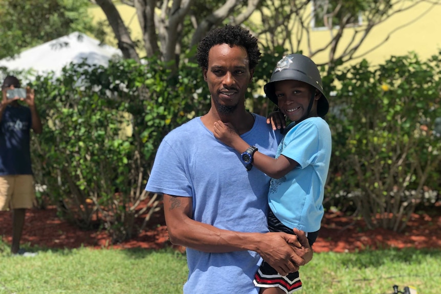 Jovan Cambridge, standing outside, holds his young son in his arms.