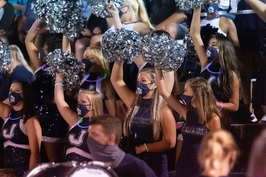 A group of cheer leaders wear masks as they wave pompoms.