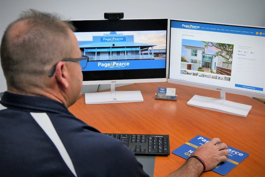 A man sits at a desk staring at two computer screens showing a house listing on a real estate website.