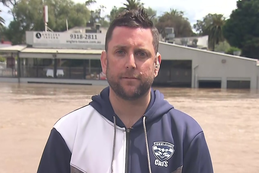 A man wearing a grey top stands in front of a flooded building.