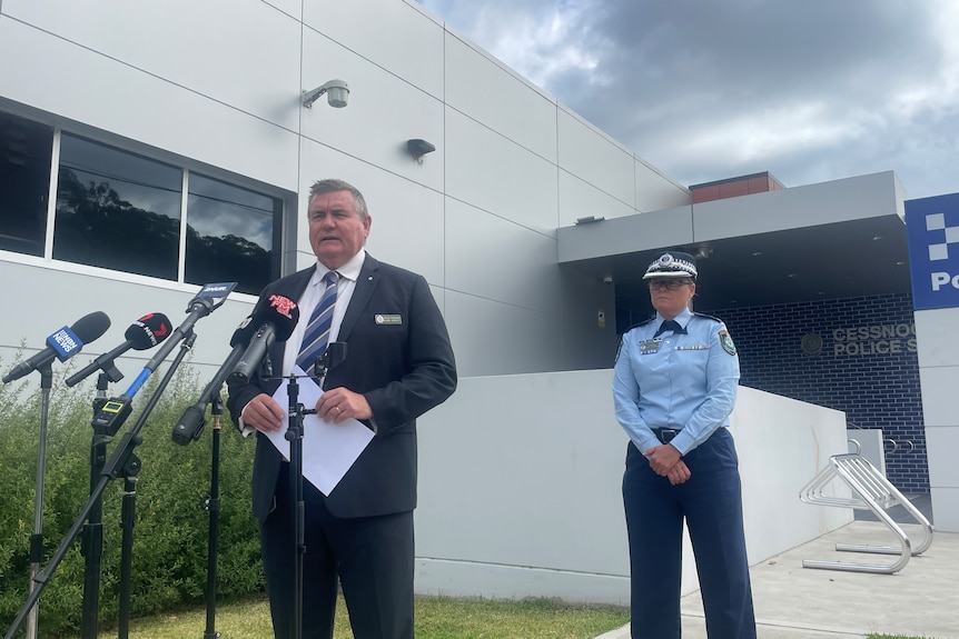 A man wearing a suit standing in front of media microphones at a press conference with a female police officer behind him