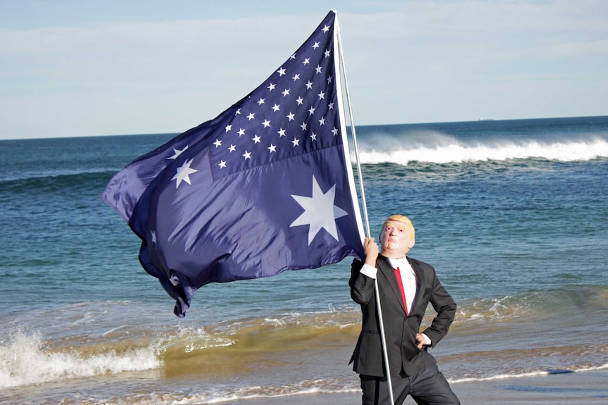 A Donald Trump impersonator holding a combined US/Australian flag on the beach.