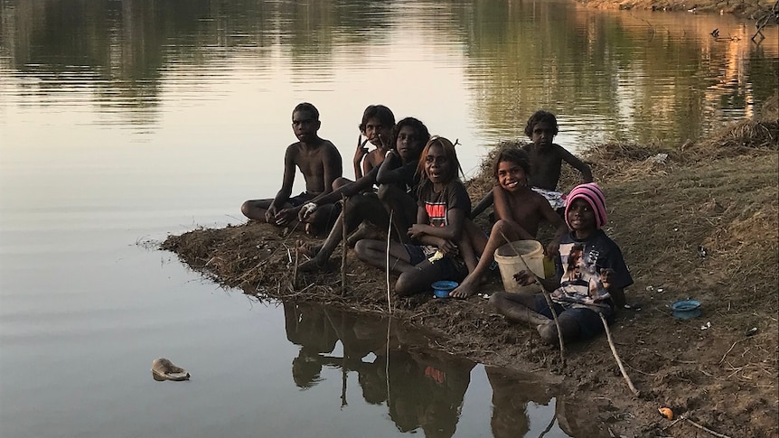 Children smile at the camera as they sit by a river