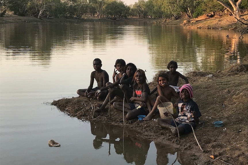 Children smile at the camera as they sit by a river