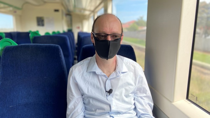 A man sits on an empty train carriage, looking out the window. He wears a white shirt, black face mask and glasses.