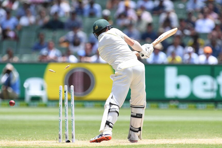Australian batsman Travis Head is seen from behind, swinging his bat as the ball hits his stumps during a Test at the MCG.