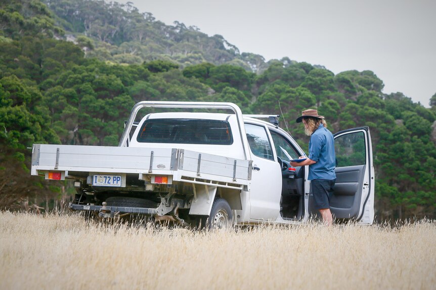 Man wearing a hat standing by a ute, with long grass in foreground and native trees behind.