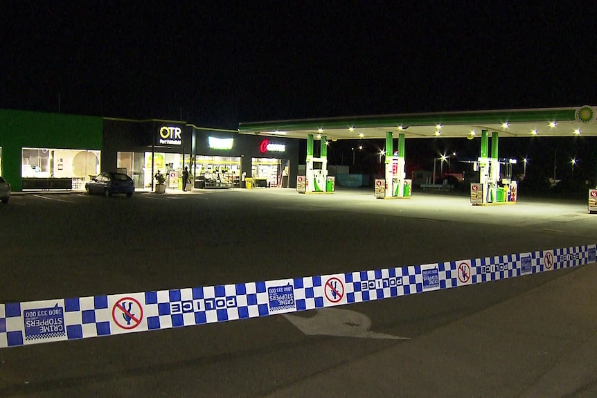 Police tape stretching across the foreground with a service station in the background at night